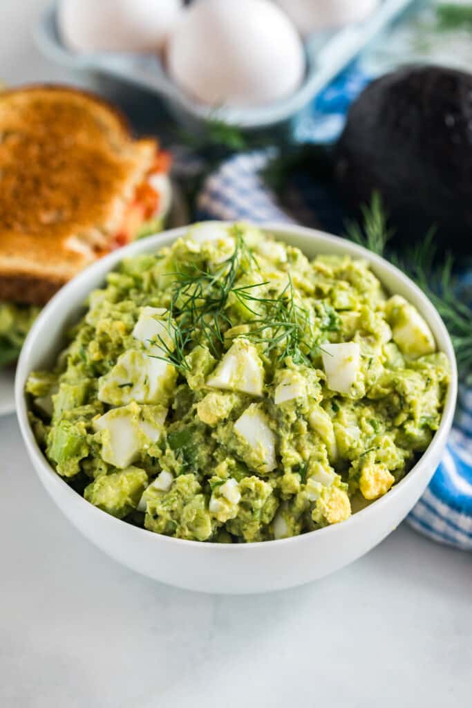A bowl of egg salad with avocado and some fresh dill on top.