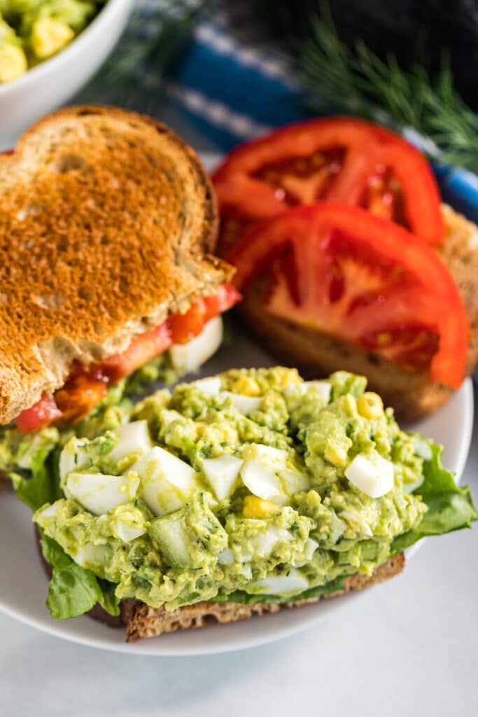 An avocado egg salad sandwich on a plate with the top of the sandwich off so you can see the egg salad