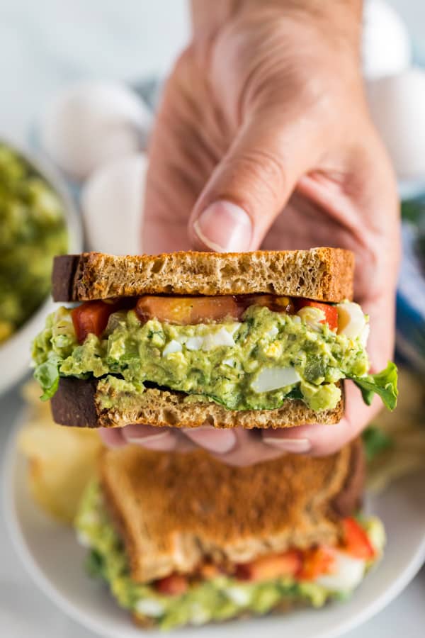 A hand holding an avocado egg salad sandwich