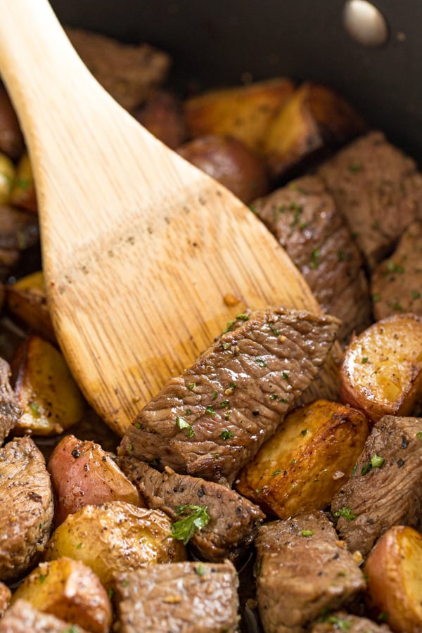 A wooden spoon in a pan of steak and potatoes.