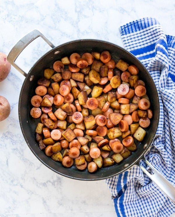 An overhead image of a saute pan full of fried potatoes and hot dogs with a blue and white checked towel next to it as well as whole red potatoes.