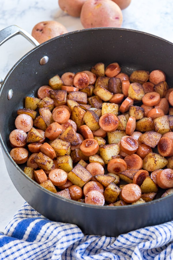 A side shot of a saute pan filed with hot dogs and potatoes with whole red potatoes behid it and a blue and white checked towel in front of it.