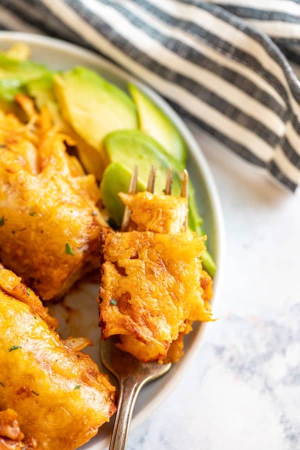 A close up image of a fork with a bite of enchiladas on with with other enchiladas on the side and sliced avocado.