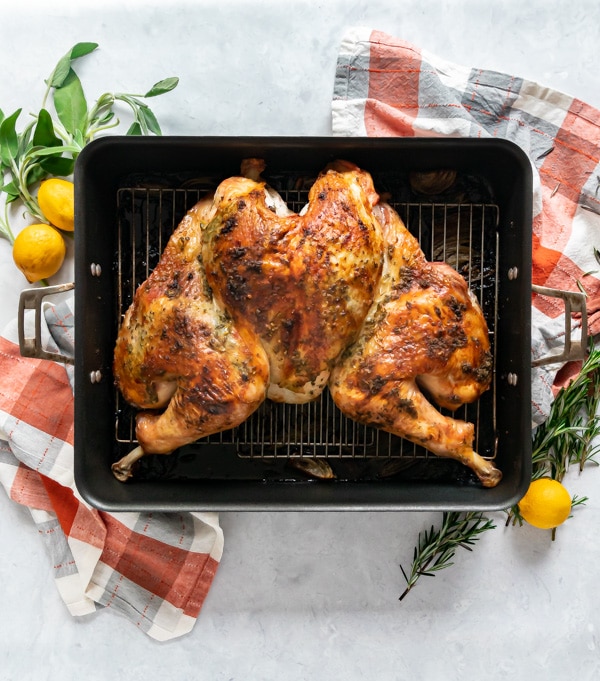 An overhead image of a roasted spatchcocked turkey in a roasting pan surrounded by fresh herbs and lemons and orange and grey plaid napkins.