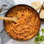 A square overhead image of a pan of penne vodka with a spoon in it surrounded by bread, basil and a striped towel.