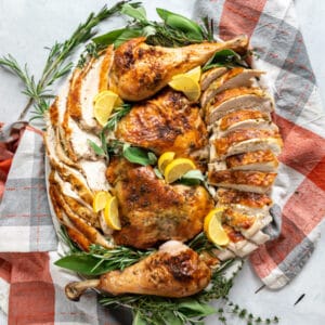 An overhead image of a carved turkey arranged on a serving platter with fresh herbs and lemons surrounding it.