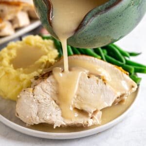 A square image of gravy being poured onto turkey breast slices on a thanksgiving plate.