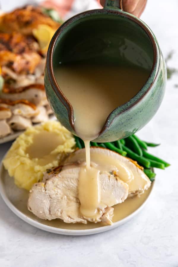 A gravy boat pouring gravy on turkey breast with green beans and mashed potatoes on the plate and a platter of turkey in the background.