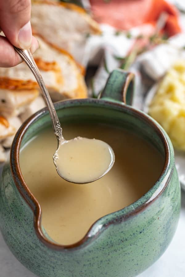 A ladle full of turkey gravy being lifted from a gravy boat full of turkey gravy with a platter of turkey in the background.
