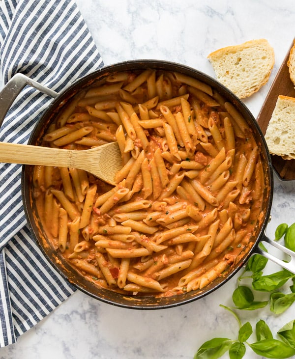 An overhead image of a pan of penne vodka with a spoon in it surrounded by bread, basil and a striped towel.