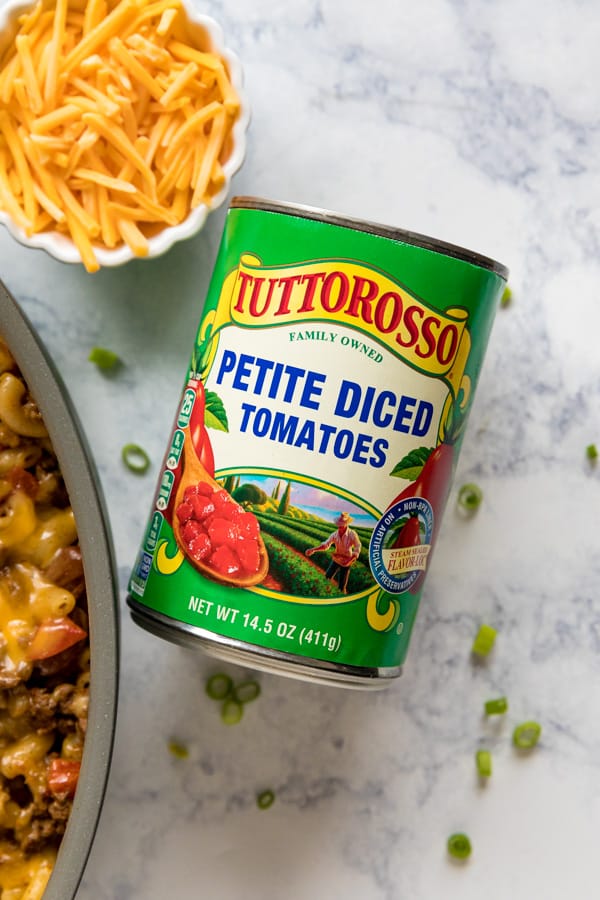 An overhead image of a can of Tuttorosso petite diced tomatoes next to the side of a pan of chili mac, a white bowl of shredded cheese and chopped scallions around it.