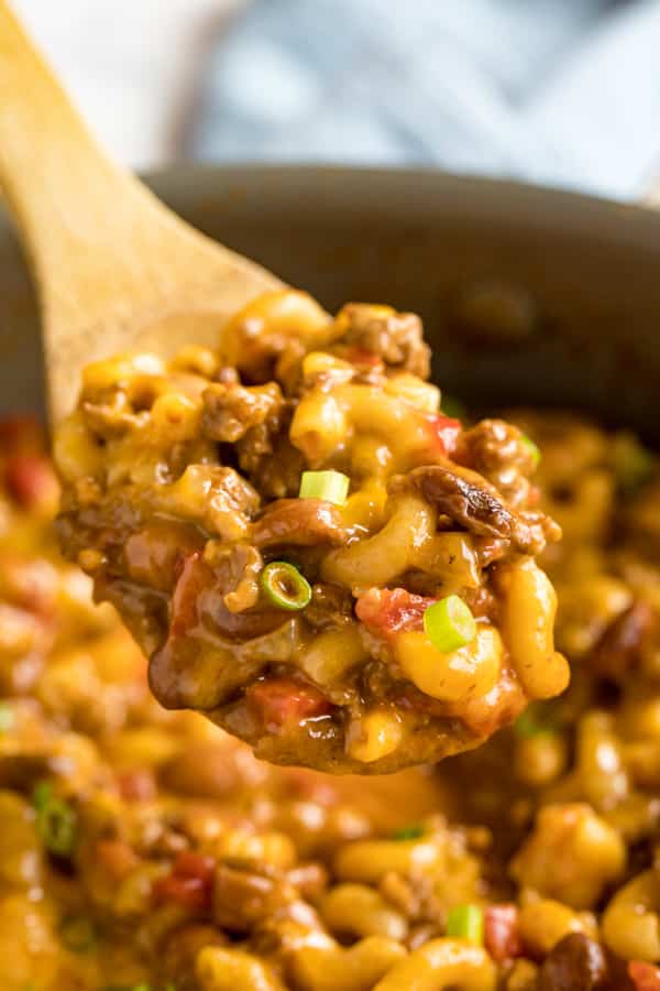 A wooden spoon holding a scoop of chili mac with the rest of the pan in the background.