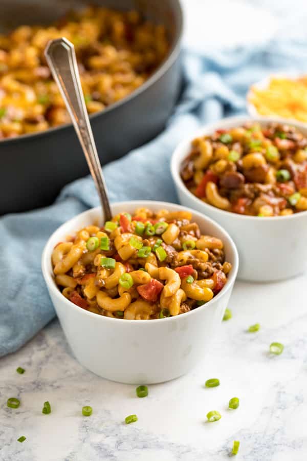 A white bowl of chili mac with a fork in it and another bowl behind it as well as the pan and a blue towel.