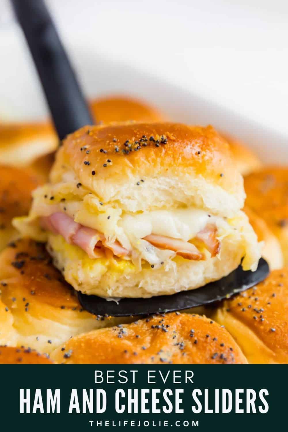 A ham and cheese slider on a spatula on top of a pan of other sliders.