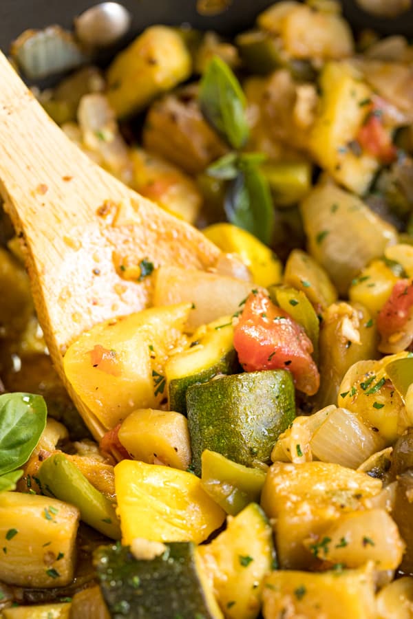 A close up image of a wooden spoon in a pan of Ratatouille focusing on the chunks of squash and eggplant.