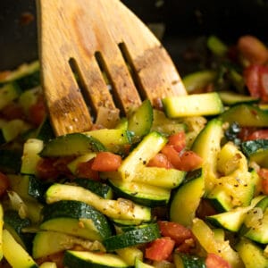 A square image of Sautéed Zucchini and Tomatoes in a sauté pan with a wooden spoon in it.