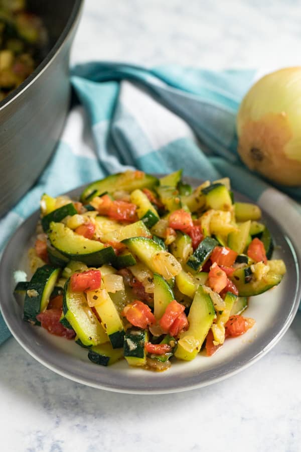 A plate of Sautéed Zucchini and Tomatoes with an onion and blue and white checked napkin behind it.