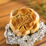 A close up image of a bulb of roasted garlic revealing it inside on top of some tin foil on a cutting board.