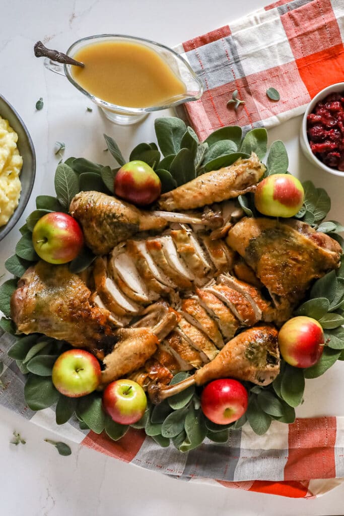 An overhead image of a carved up turkey on a platter with sage and apples with grav on the side of it and orange and grey napkins..