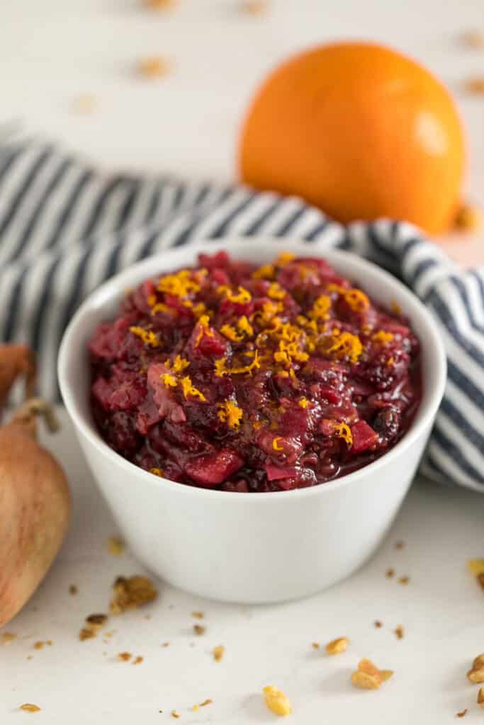 A white bowl of chutney with cranberries and orange zest on top with a striped towel and an orange behind it.