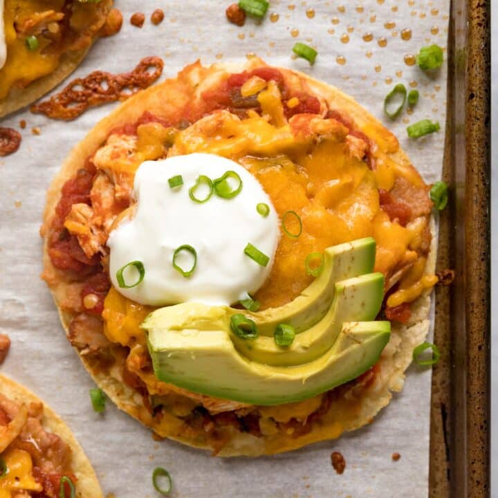 An overhead image focusing on a tostada on a parchment-lined pan topped with sour cream, sliced avocado and green onion.