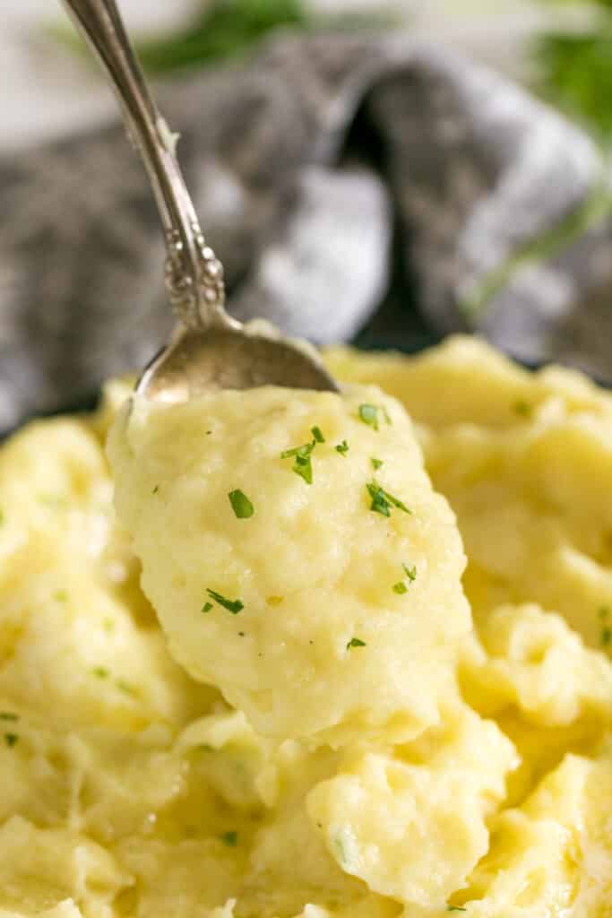 A spoon holding a scoop of mashed potatoes with a bowl of them behind it and a grey napkin.