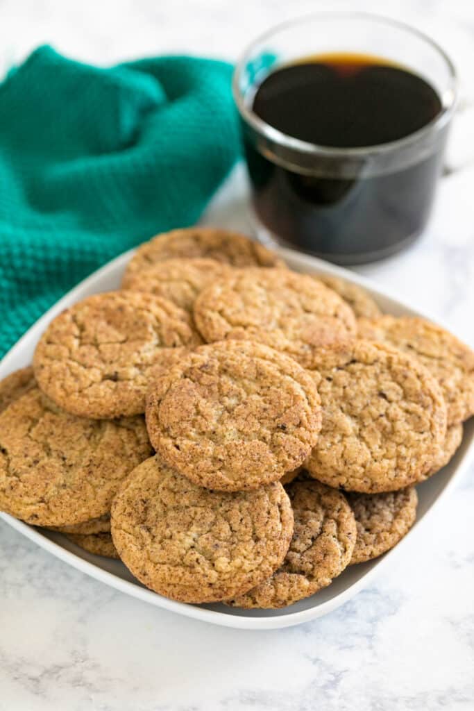 Cookies piled on a square plate with coffee and a green towel in the background.