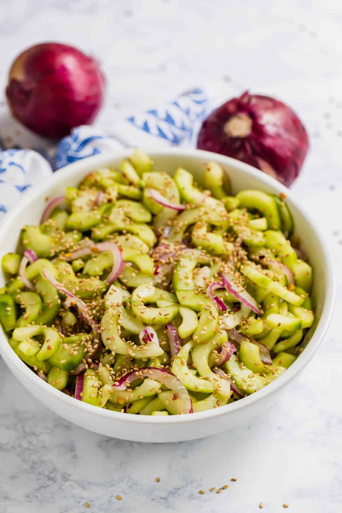 A bowl of cucumber salad with a blow towel and red onions behind it.