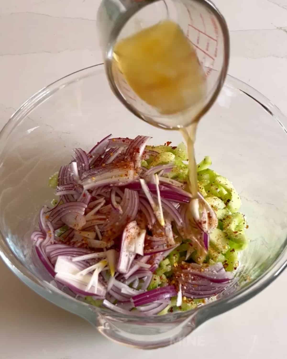 Dressing being poured over cucumber salad in a mixing bowl.