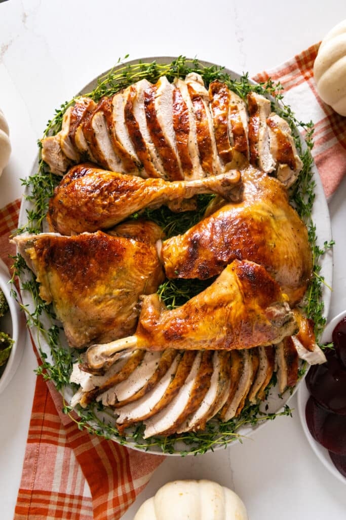 An overhead image of sliced turkey on a platter surrounded by fresh herbs in the middle of a Thanksgiving table.