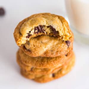 Chocolate chip cookies stacked with a bite in the top cookie exposing melted chocolate chips and stacked next to a glass of milk.