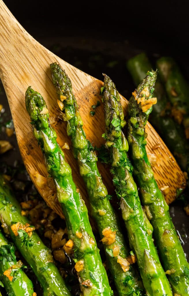 Four sautéed asparagus spears held up on a wooden spatula in a pan.