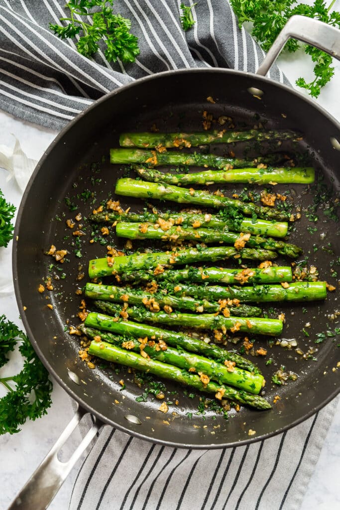 An overhead image of a black sauté pan with perfectly pan seared asparagus with garlic and parsley around it.