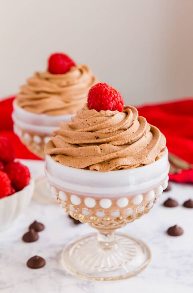 An image of a chocolate mousse cup with another in the back; both are topped with raspberries and have some chocolate chips around them.