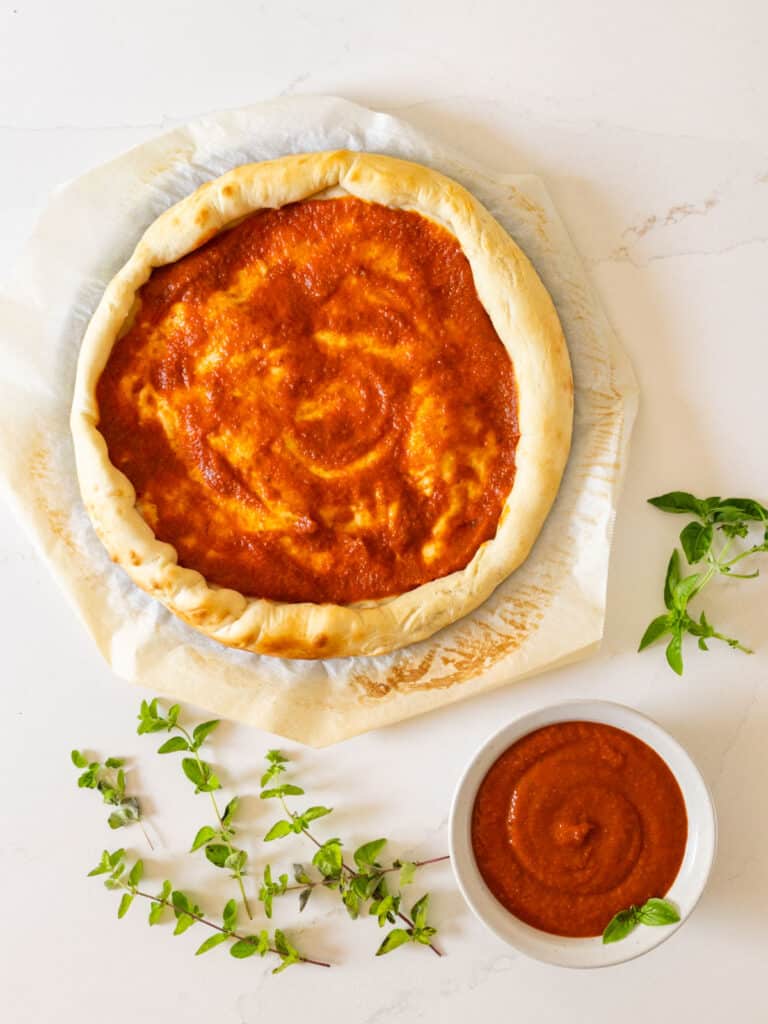 An overhead image of pizza crust with sauce on it and a bowl of sauce with sprigs of fresh basil and oregano around it.