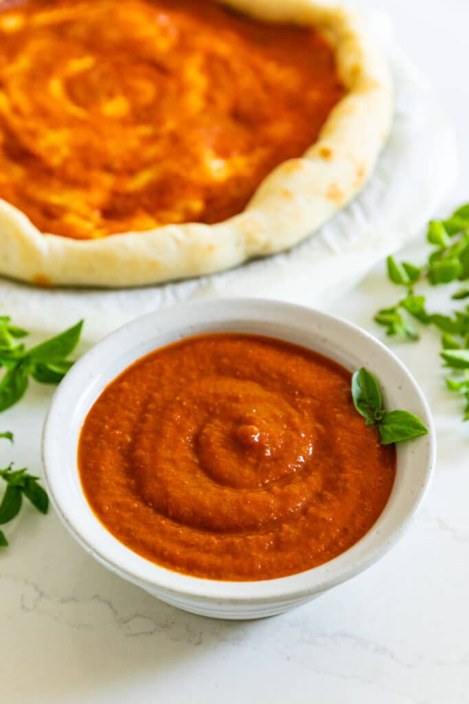 A bowl of pizza sauce with a sprig of basil in it and pizza dough with sauce on it in the background.