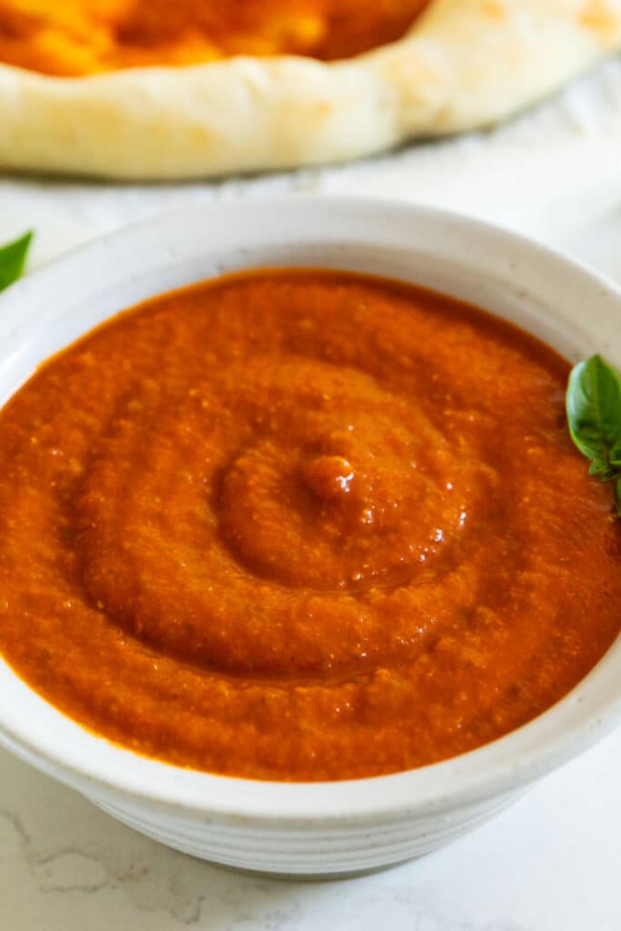 A close up image of a bowl of pizza sauce with a sprig of basil in it.