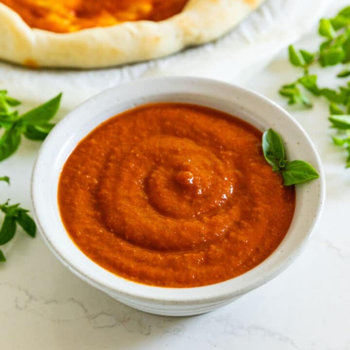 A bowl of pizza sauce with a sprig of basil in it and pizza dough with sauce on it in the background.