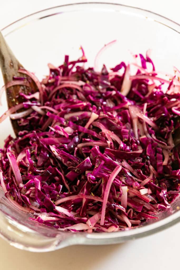 A close up of a glass mixing bowl full of cabbage salad with wooden salad spoons in it.