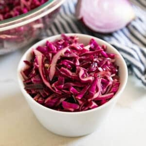 A white bowl of red cabbage salad with a large glass bowl of it and a chunk of red onion in the background.