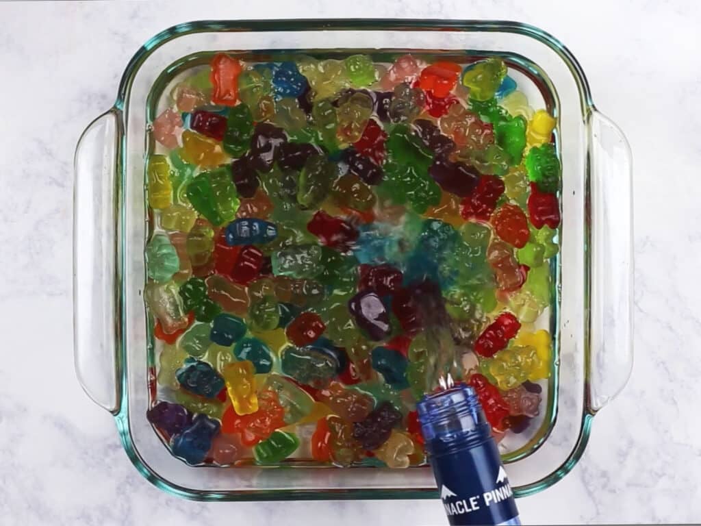 Vodka being poured over the gummy bears in the glass container.
