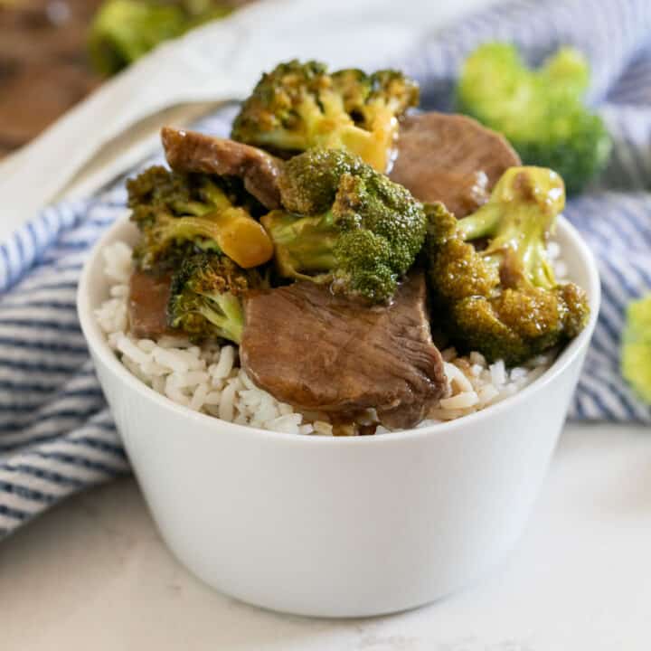A white bowl with white rice and beef and broccoli on top with a blue and white striped napkin next to it and the sheet pan in the background.
