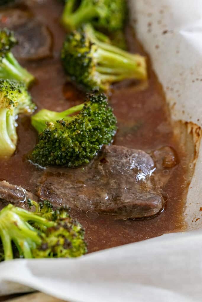 A close up image of some sliced steak and broccoli with brown sauce on the side of a parchment lined sheet pan.