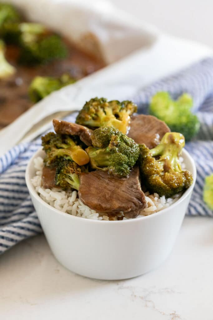 A white bowl with white rice and beef and broccoli on top with a blue and white striped napkin next to it and the sheet pan in the background.