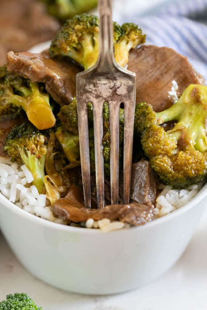 A bowl of beef and broccoli over rice with a fork piercing into a piece of beef.