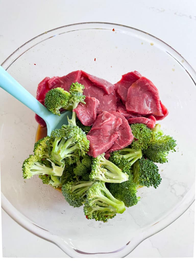 Sliced steak and raw broccoli in a clear bowl with the marinade.