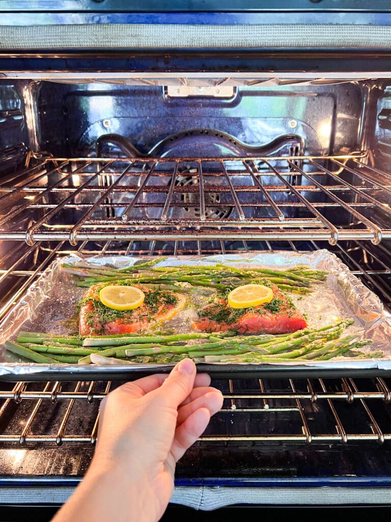 A hand placing the sheet pan of salmon and asparagus into the oven.