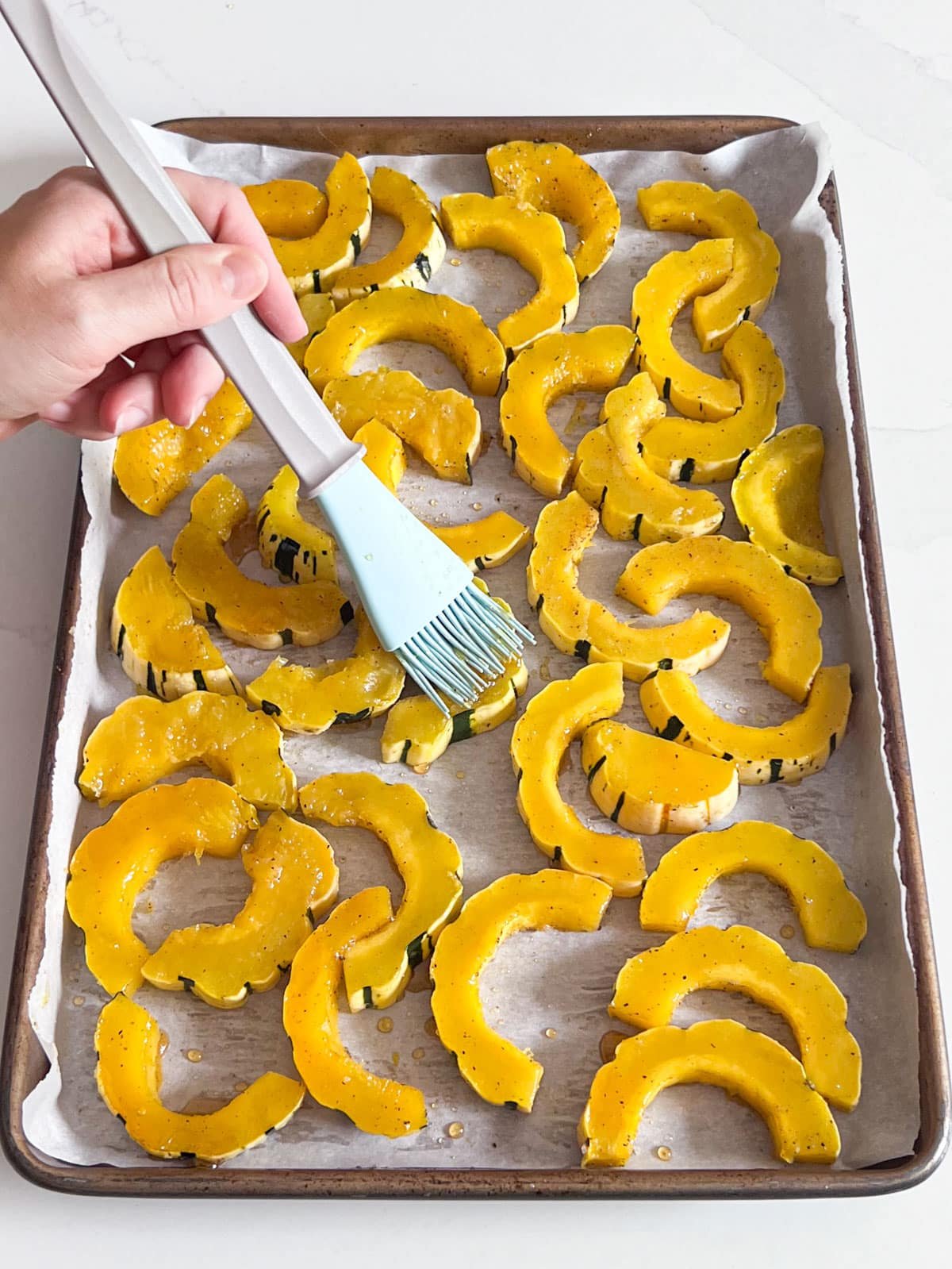 A hand brushing hones on slices of delicata squash on a parchment-lined pan.