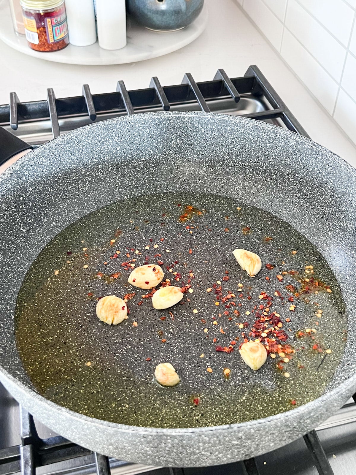 A skillet on a stove with oil, garlic and chili flakes cooking in it.