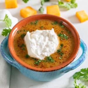 A bowl of squash soup with burrata and cilantro on top and chunks of squash and cilantro in the background.